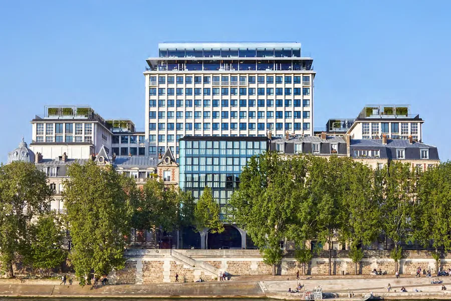 L’ancienne Cité administrative de Paris vue depuis la Seine : la tour de 1964 coiffée d’un niveau vitré, un socle de verre en pied, entre les immeubles haussmanniens et les arbres du quai.
